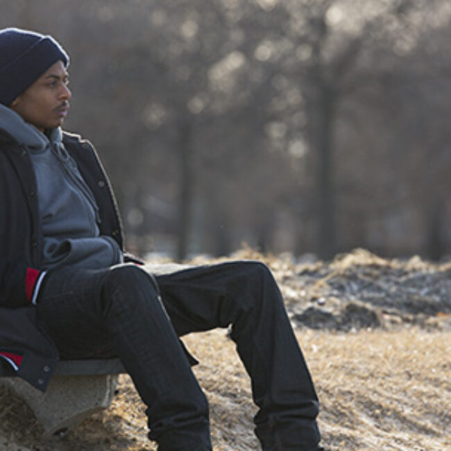 Young African American man sits on a park bench in winter clothing.