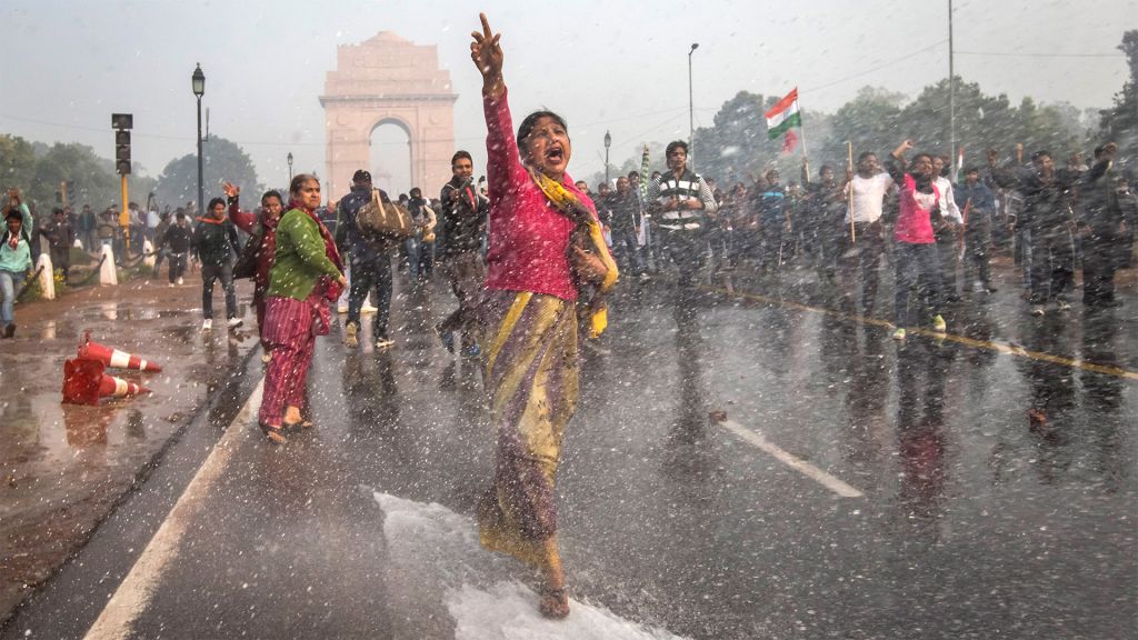 A woman marches down a Delhi street shouting in protest as water cannons shower the scene.