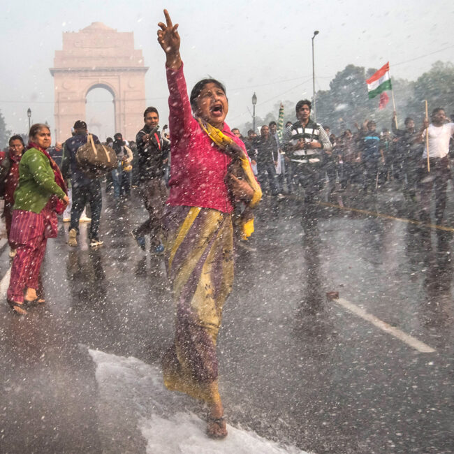 A woman marches down a Delhi street shouting in protest as water cannons shower the scene.