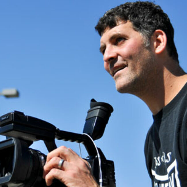 Filmmaker Brad Lichtenstein smiles while framing a shot with a video camera.