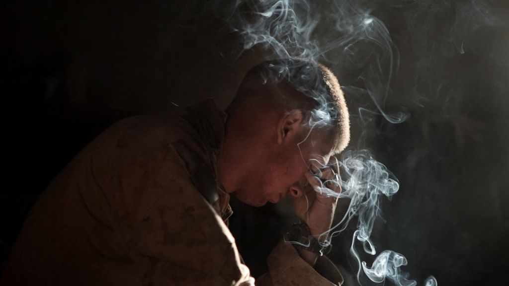 A solider holds his head in his hand with cigarette smoke curling in the air.