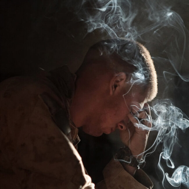 A solider holds his head in his hand with cigarette smoke curling in the air.