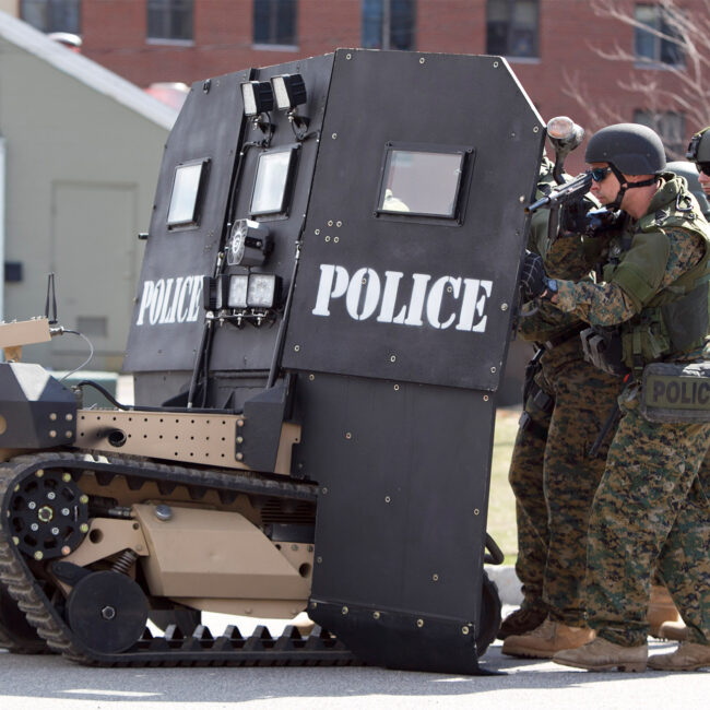 Police in military fatigues huddle behind a SWAT robot