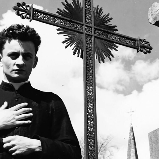 Still of priest standing in front of outdoor cross in Robert Bresson's 1951 film Diary of a Country Priest