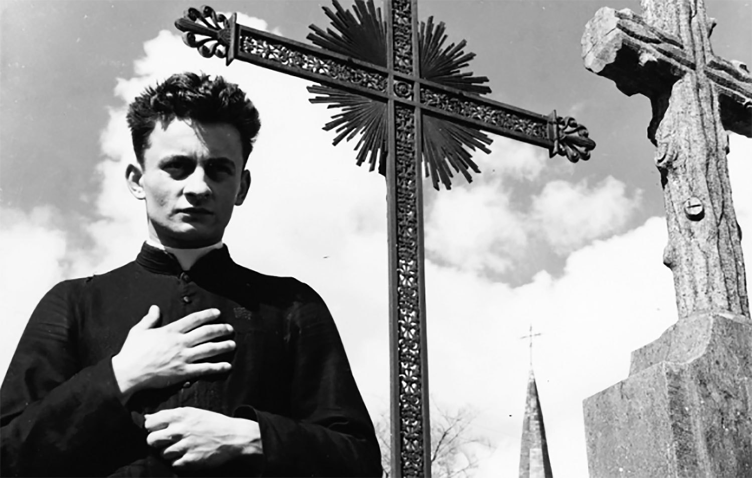 Still of priest standing in front of outdoor cross in Robert Bresson's 1951 film Diary of a Country Priest