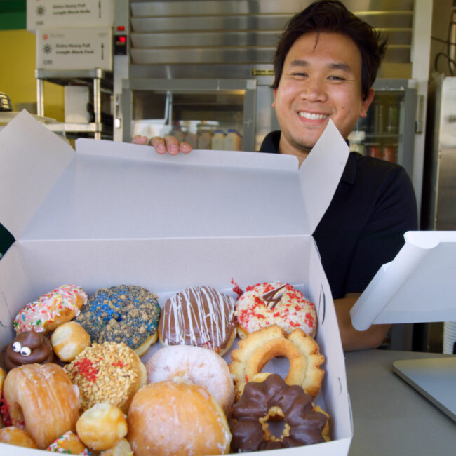 A smiling Cambodian American donut clerk offers up a box full of donuts in Southern California