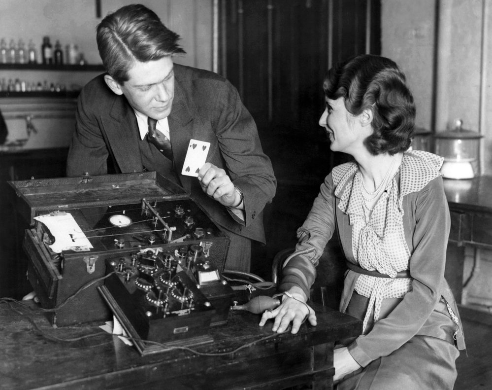 A blonde man holding a playing card leans over a lie detector machine with a woman looking on.