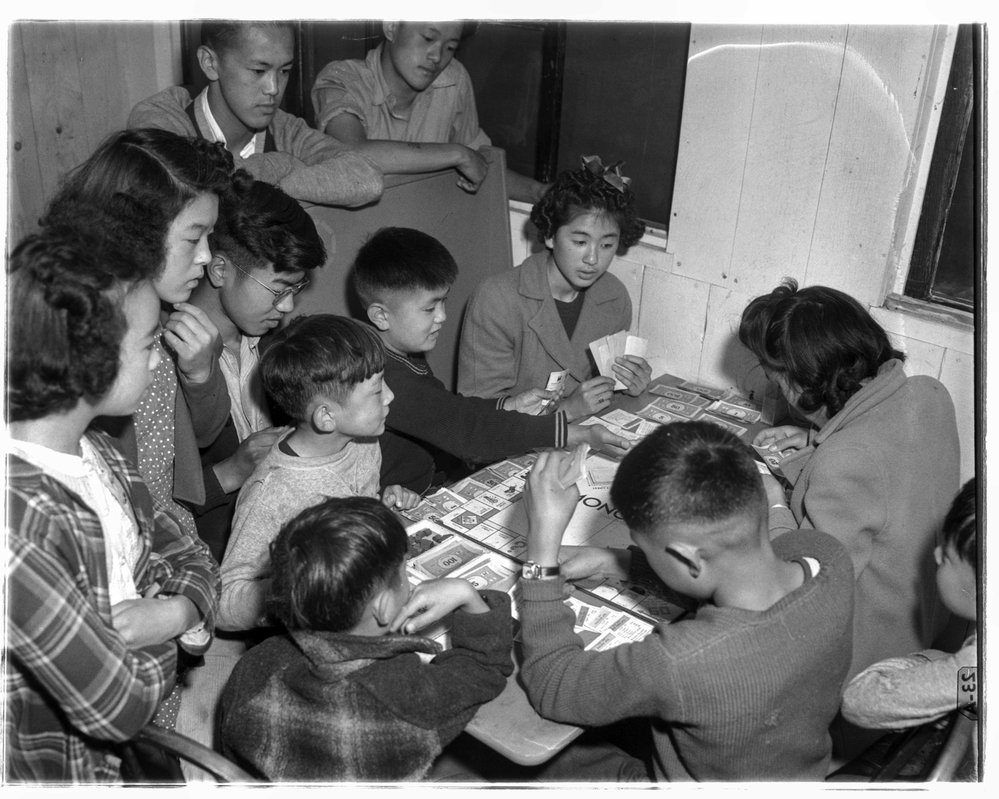 Six Japanese American children huddle around a table playing Monopoly while six others look on.