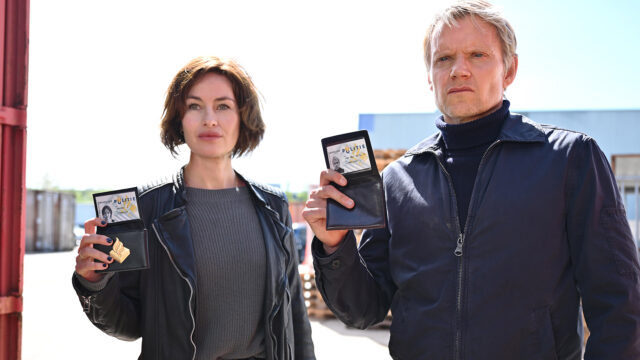 Maimie McCoy as Lucienne Hassell and Marc Warren as Piet Van der Valk holding up badges in Van der Valk Season 3