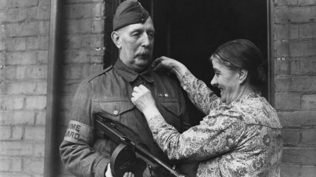 Black and white photo of an older British man serving in the Home Guard during WW2, with his wife adjusting his uniform.