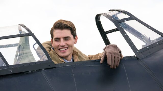 Actor Gregg Sulkin as RAF pilot David, sitting in the cockpit of his fighter plane, smiling broadly.